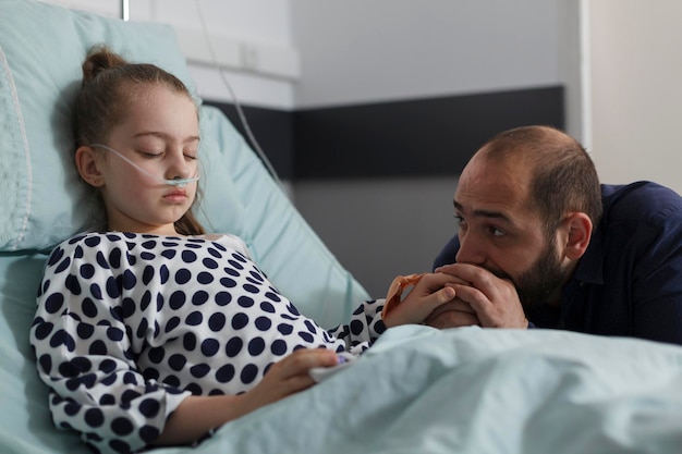 体調不良の子供が寝ている間、不安な親が医療施設内の彼女のそばに座っている.病院の小児科病棟内の患者のベッドで休んでいる病気の少女の隣に座っている心配している父親。