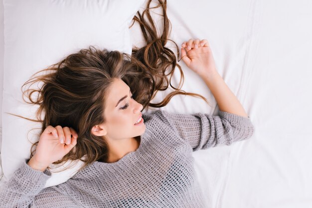 Up portrait cheerful girl with long brunette hair relaxing on white bad. Good morning, positive emotions, smiling with closed eyes, chilling at home, dreaming. Place for text.