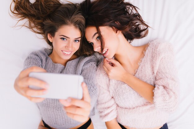 보기 흰색 침대에 selfie 초상화를 만드는 두 매력적인 즐거운 젊은 여성. 따뜻한 니트 울른 스웨터를 입은 예쁜 소녀들의 집에서 쿨링. 재미 있고, 웃고, 좋은 아침.