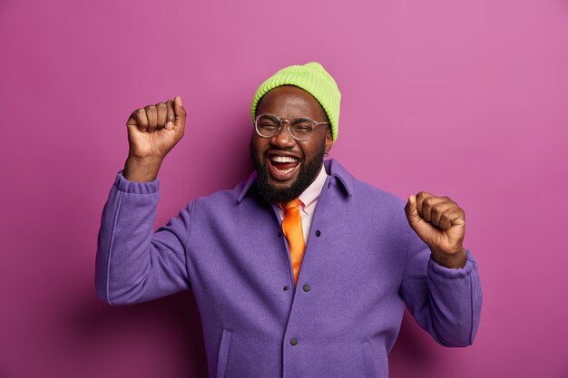 Upbeat carefree black bearded man dances with clenched fists, has carefree mood, cheers and moves actively, rejoices nice event, wears fashionable bright clothes, enjoys pleasant music and tunes.