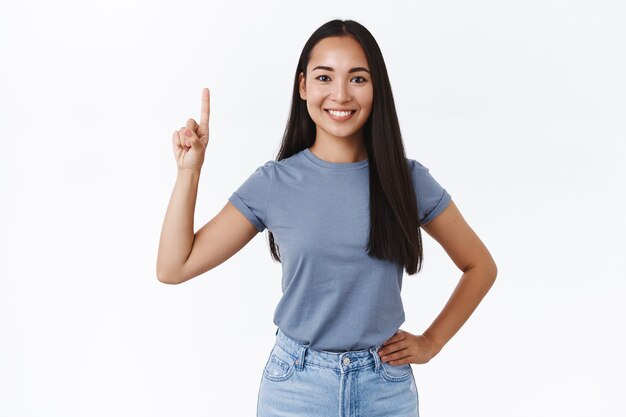 カジュアルなTシャツを着た明るい、のんきな笑顔のアジアの女性ブルネット、前向きな姿勢で手を腰に当て、指を上に向け、1つを見せ、ニヤリと、何を買うかアドバイスを与え、製品を宣伝する