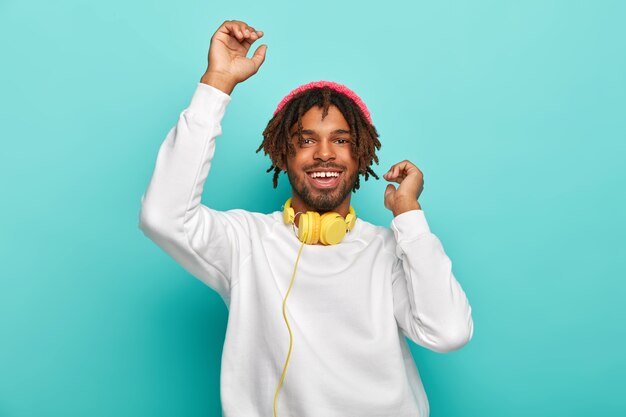 Upbeat carefree mixed race guy dances with hands up, listens awesome music track via headphones, has dreads, wears white comfortable jumper and stylish hat