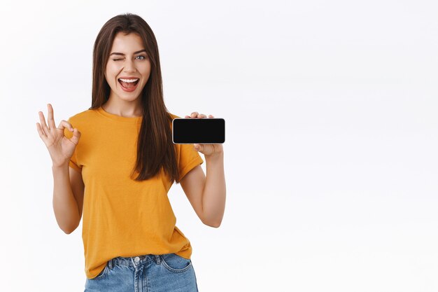 Upbeat, cheerful pretty brunette woman in yellow t-shirt holding smartphone horizontally, make okay, good excellent gesture and winking, smiling satisfied promote application or mobile game