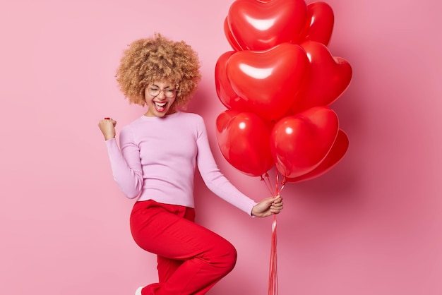 밝은 곱슬머리 여성은 발렌타인 데이를 축하하는 기쁨으로 주먹을 꽉 쥐고 하트 모양의 풍선을 들고 분홍색 배경 휴일과 축하에 격리된 캐주얼 점퍼와 바지를 입습니다.