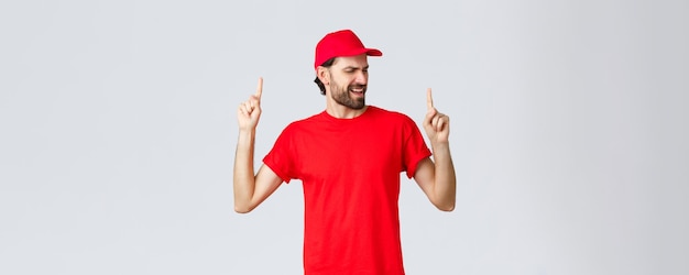 Upbeat handsome bearded delivery guy spending break relaxing employee in red uniform cap and tshirt singing song pointing fingers up carefree close eyes standing grey background