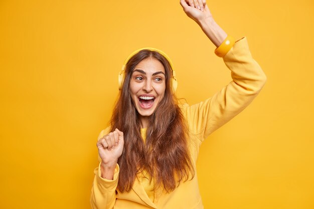 Upbeat happy long haired woman makes triumphing dance raises arms enjoys favorite music listens songs via headphones foolishes around isolated over yellow wall. Lifestyle and hobby concept