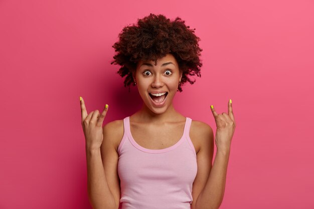 Upbeat positive ethnic woman makes rock n roll goat gesture, being real devoted rocker, attends wild party, has joyful expression, dances and has fun, isolated over pink wall, being crazy