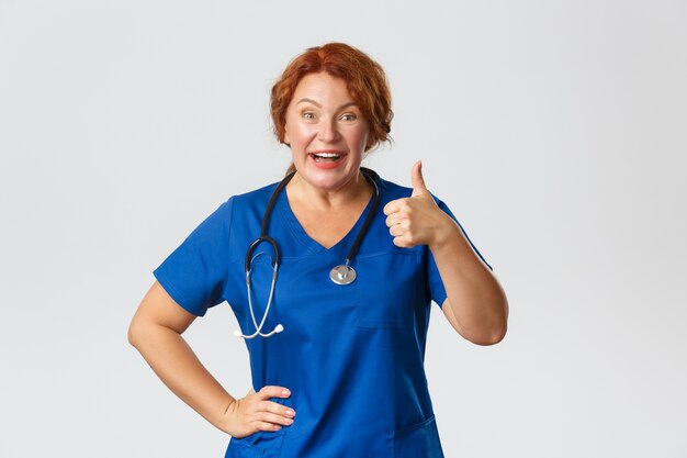 Upbeat redhead middle-aged doctor recommend clinic offer or covid-19 test discount, showing thumbs-up and smiling, approve choice, 