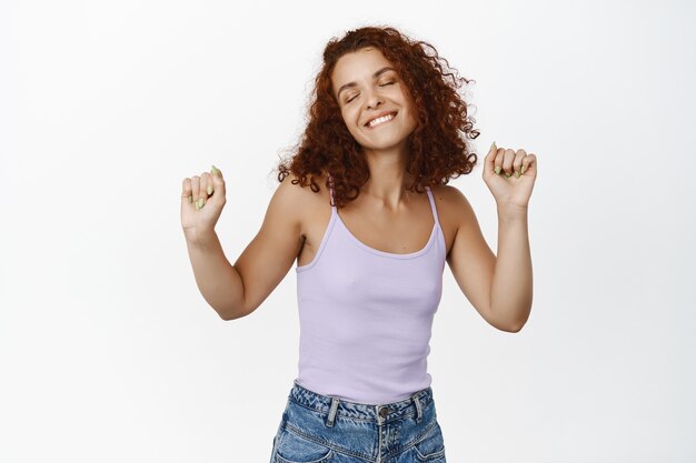 Upbeat redhead woman dancing, raising hands up and smiling carefree, partying, enjoying music with relaxed face expression and eyes closed on white