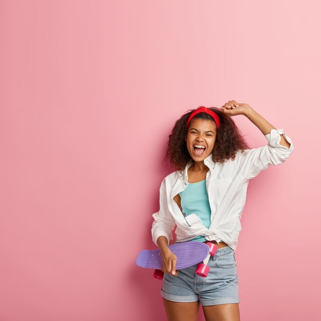 Upbeat young Afro American woman raises hand, feels pleased, wears white shirt and denim shorts, exclaims with happiness, keeps mouth opened, carries purple longboard, models against pink wall