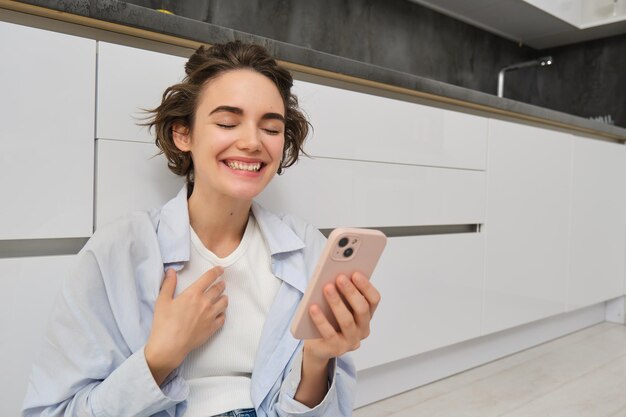 Upbeat young woman talking on mobile phone video chats and laughs points at herself uses smartphone