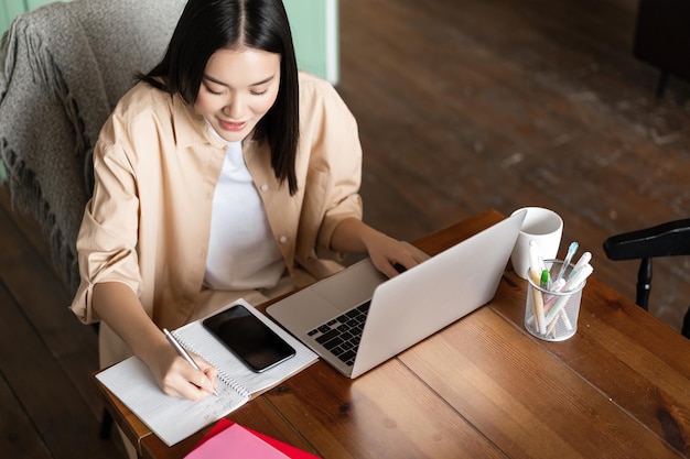 Upper angle of asian girl working indoors woman studying remotely writing down notes doing homework ...