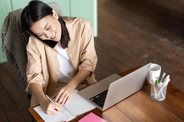 Верхний угол снимок азиатской женщины, работающей с ноутбуком, отвечающей на телефонный звонок и записывающей в блокноте g ...