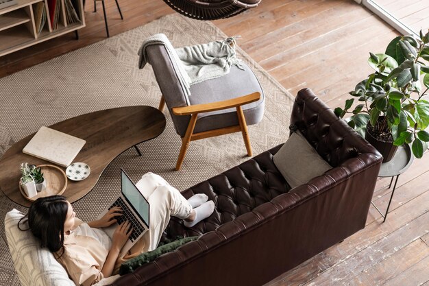 Upper angle shot of young asian woman laying on couch and using laptop working on computer or studyi...