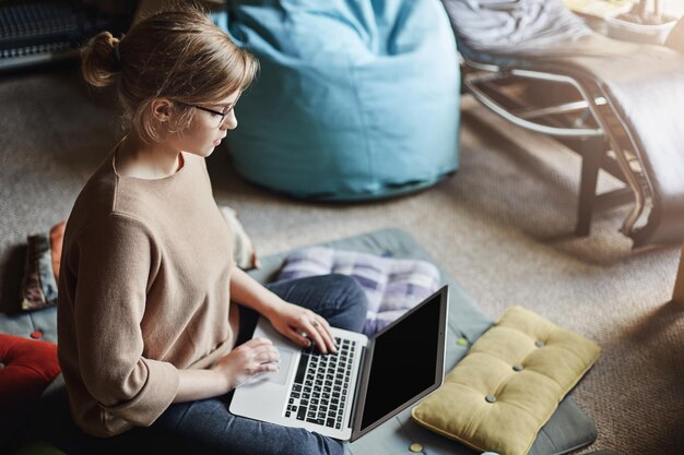 Снимок сверху: решительная привлекательная женщина в уютном наряде сидит на полу с ноутбуком на коленях и смотрит в экран, собирая данные для университетского проекта, усердно работая без перерыва