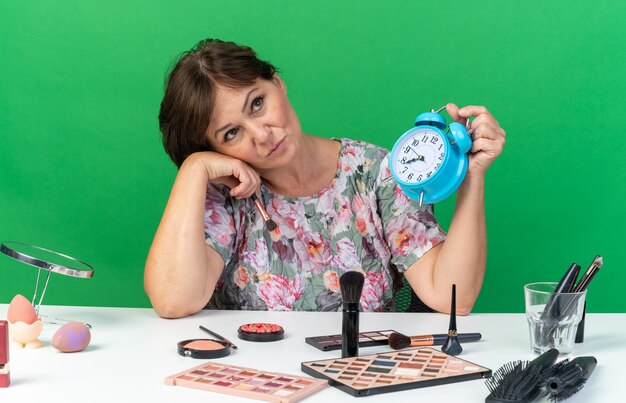 Upset adult caucasian woman sitting at table with makeup tools holding alarm clock and makeup brush looking up isolated on green wall with copy space