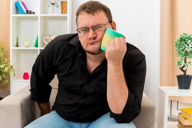 Upset adult slavic man in optical glasses sits on armchair holding and putting cup on face