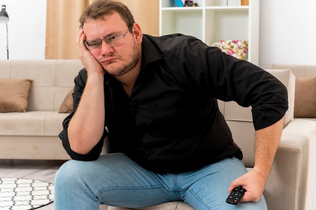 Upset adult slavic man in optical glasses sits on armchair putting hand on face