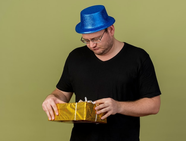 Sconvolto uomo slavo adulto in occhiali ottici che indossa un cappello da festa blu tiene e guarda la confezione regalo