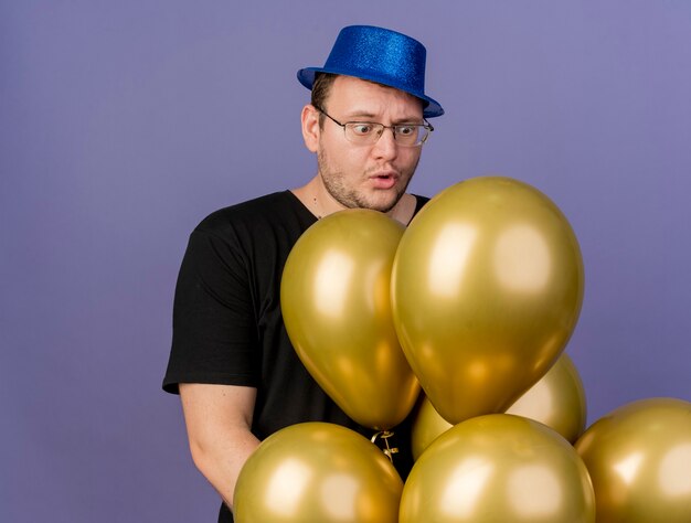 Upset adult slavic man in optical glasses wearing blue party hat holds and looks at helium balloons 