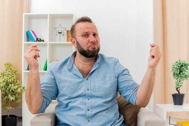 Upset adult slavic man sits on armchair gesturing money hand sign with two hands looking at camera inside the living room