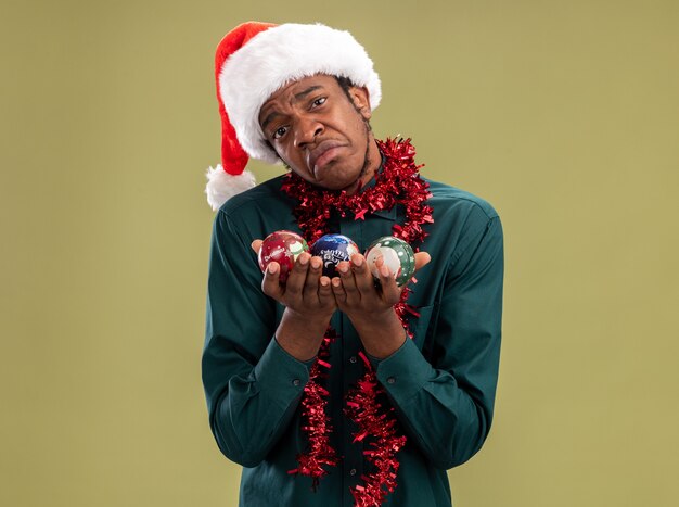 Upset african american man in santa hat with garland holding christmas balls looking at camera with sad expression pursing lips standing over green background