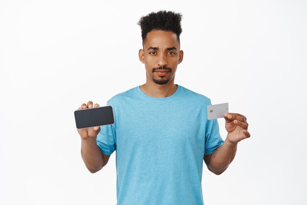 Upset african american man showing horizontal smartphone screen credit card disappointed with banking app no money bank account standing displeased against white background