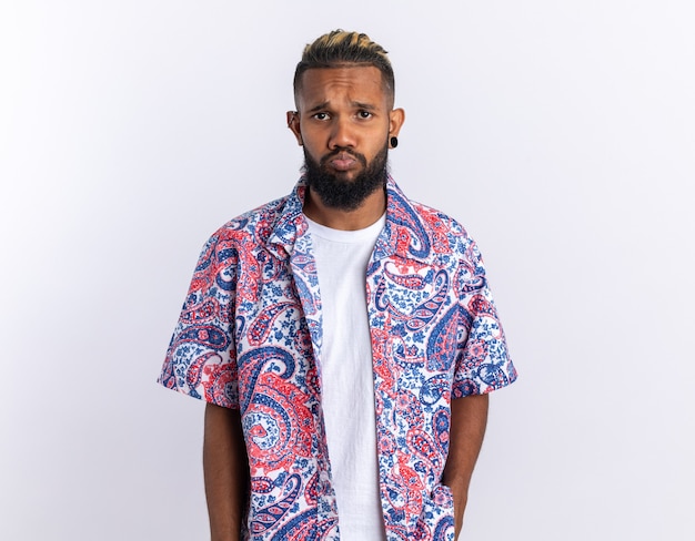 Upset african american young man in colorful shirt looking at camera with sad expression standing over white background