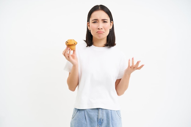 カップケーキを持って動揺し、ジャンクな甘い食べ物を食べたいと不平を言うアジア人女性が白い背景の上に立っている