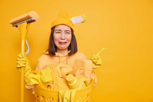 Расстроенная азиатская женщина-уборщик чувствует себя грустной и усталой, использует швабру для уборки дома, позирует возле бассейна с небрежно одетым бельем, указывает в верхнем правом углу, изолированном на желтом фоне. Уборка дома