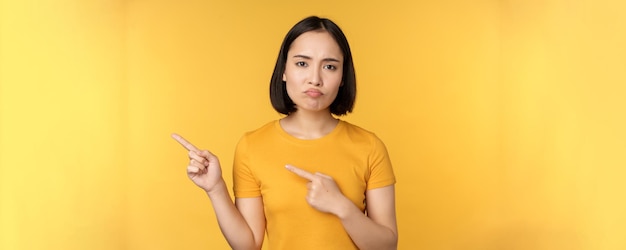 Upset asian woman pointing fingers left showing smth disappointing complaining at banner standing over yellow background