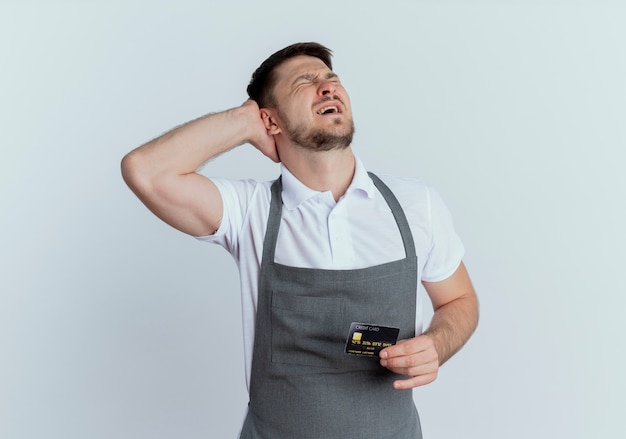 Upset barber man in apron holding credit card with closed eyes standing over white background