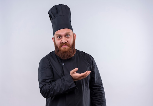 An upset bearded chef man in black uniform raising hand with surprised face on a white wall