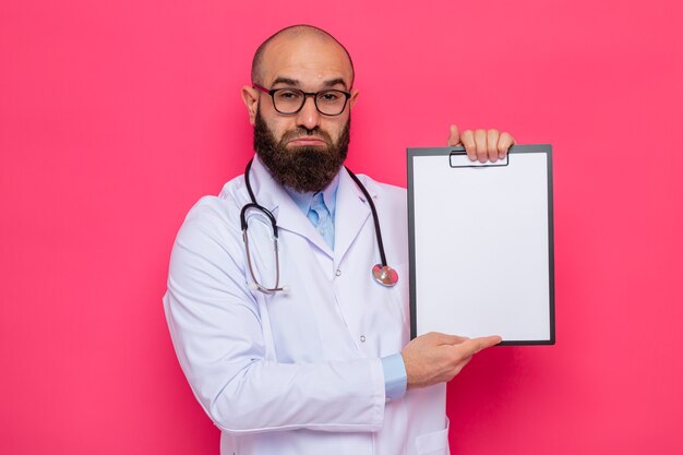 Расстроенный бородатый мужчина-врач в белом халате со стетоскопом на шее в очках, держит буфер обмена с пустыми страницами, смотрит с грустным выражением лица, поджимая губы