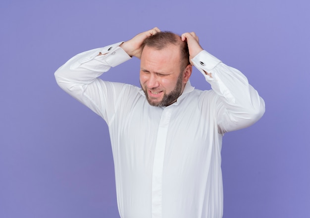 Upset bearded man wearing white shirt touching his head being frustrated standing over blue wall