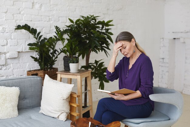 Upset beautiful 60 year old woman sitting on gray chair in living room, touching forehead and looking at open copybook on her lap, feeling frustrated because she forgot about important meeting