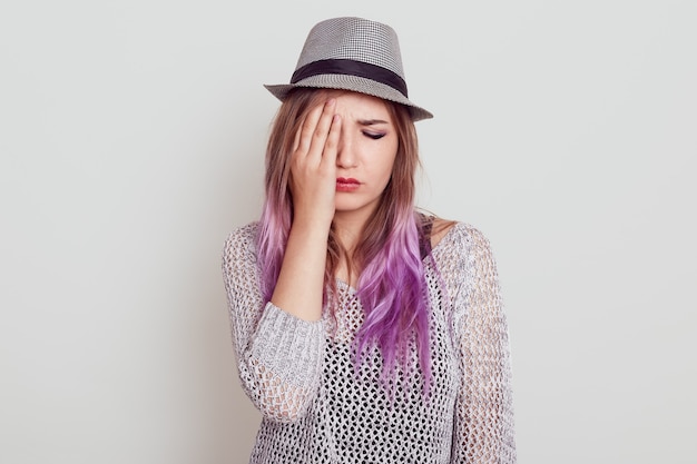 Upset beautiful woman with lilac hair wearing shirt and hat being sad, covering half of face with palm, keeps eyes closed, isolated over white wall.