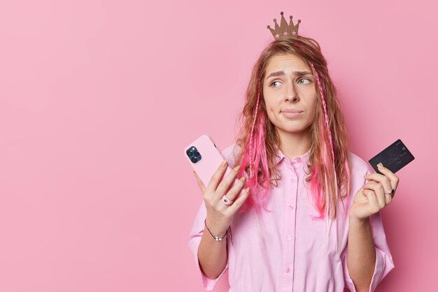 화가 난 아름다운 젊은 여성은 은행 계좌 주문에 돈이 없어 온라인에서 스마트폰과 신용 카드를 들고 분홍색 배경의 빈 공간에 고립되어 있습니다.