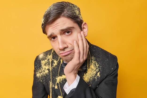 Upset birthday man feels unhappy keeps hand on cheek and looks sadly at camera smeared with serpentine spray wears formal clothes poses against yellow background feels tired after birthday party