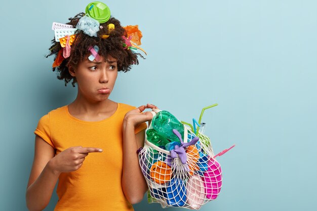 Upset black woman has curly hair, points fore finger on trash made of plastic, cleans rubbish, does environmental project, being in low spirit, wears orange t shirt, stands over blue wall