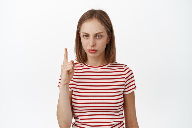 Upset blond girl pointing finger up, frowning displeased, dislike smth, express her negative opinion, unfair thing, standing against white background in t-shirt