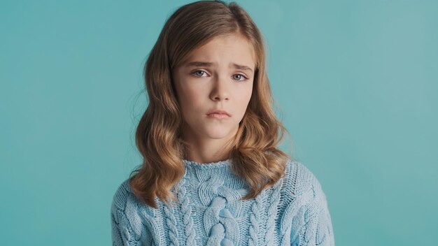 Upset blond teenage girl dressed in sweater feeling sad over blue background Offended girl