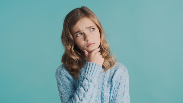 Upset blond teenager girl looking worry keeping hand on chin looking thoughtful over blue background. Face expression