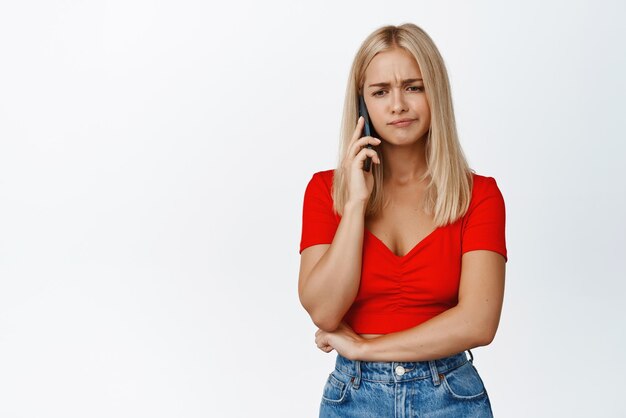 動揺しているブロンドの女性は、問題のある顔の表情の白い背景で携帯電話で話している電話で悪いニュースを受け取ります