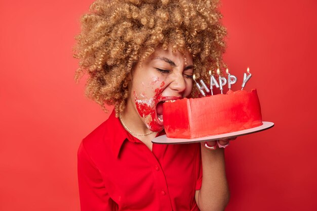 화난 금발의 곱슬머리 여성이 배고파서 맛있는 달콤한 케이크를 물고 케이크가 묻은 생일을 축하한다 선명한 붉은 배경에 홀로 포즈를 취한다 여성 모델은 축제 디저트를 실내에서 먹는다