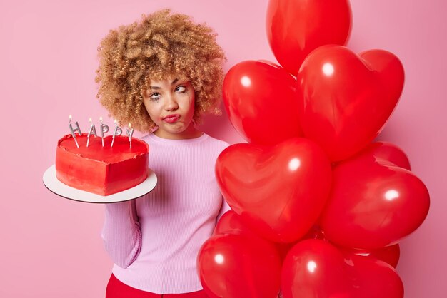 Upset bored European woman holds delicous heart shaped cake and bunch of red helium balloons has spoiled makeup wears casual jumper isolated over pink background People and holidays concept