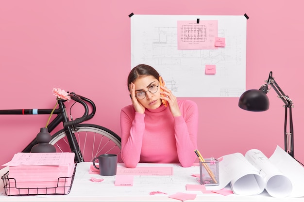 Расстроенная скучающая женщина, уставшая от работы, готовит бумажные эскизы, ведет телефонный разговор, работает над проектом архитектора, носит круглые очки, розовая водолазка, позирует на рабочем столе в окружении чертежей