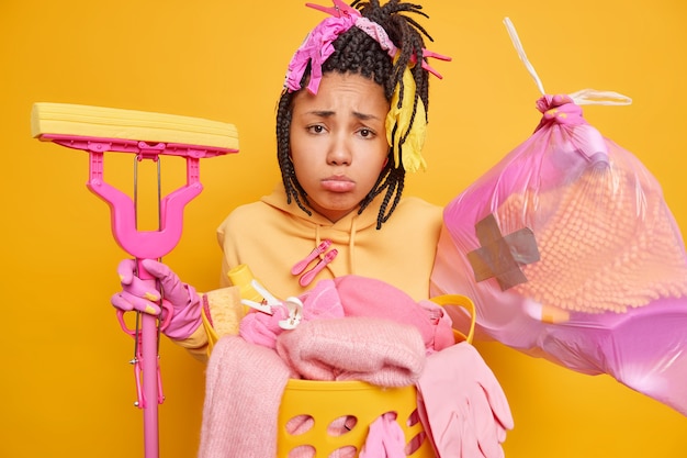 Upset bored young African American woman with dreadlocks collects rubbish in trash bag