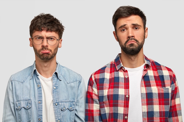 Upset brothers have unhappy expressions, frown faces in discontent, being in low spirit after watching football match, realize their favourite team loose game, isolated over white wall