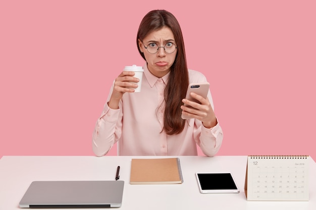 화가 갈색 머리 여성 사무실 완벽 주의자는 불행한 표정을 가지고 있고, 테이크 아웃 커피를 마시고, 휴대 전화를 들고, 둥근 안경을 착용합니다.